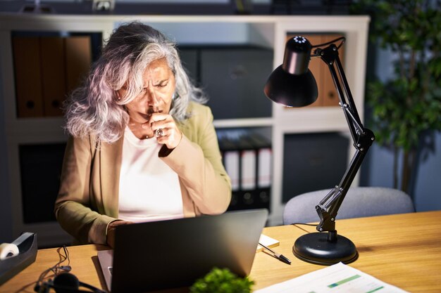 Foto donna di mezza età con i capelli grigi che lavora utilizzando il computer portatile a tarda notte sensazione di malessere e tosse come sintomo di raffreddore o bronchite concetto di assistenza sanitaria