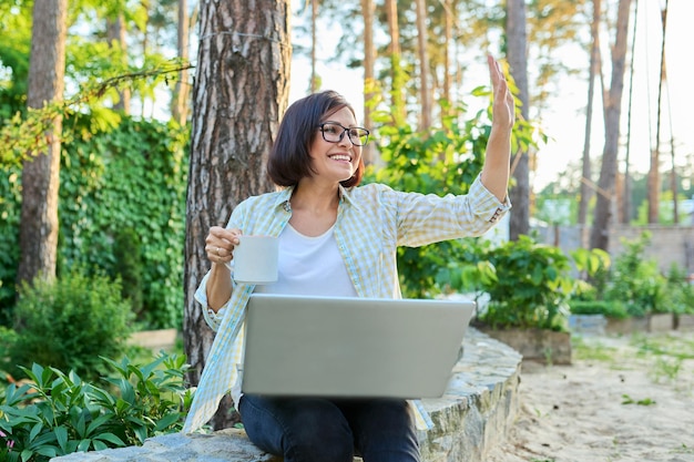 Женщина средних лет с чашкой ноутбука отдыхает на заднем дворе