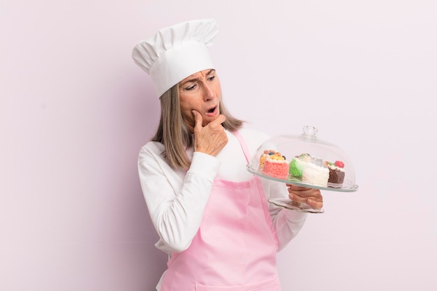 Middle age woman with mouth and eyes wide open and hand on chin baker and cakes concept