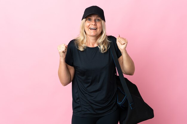 Middle age woman with sport bag isolated on pink wall celebrating a victory in winner position