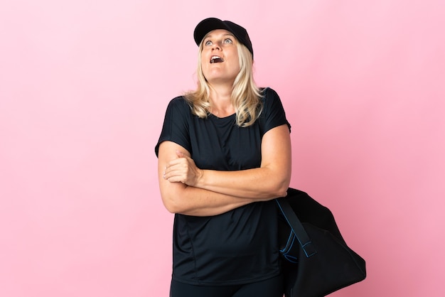 Middle age woman with sport bag isolated on pink wall looking up and with surprised expression