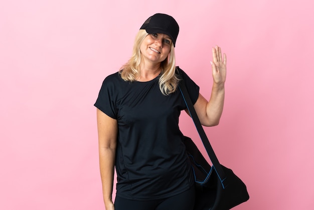 Middle age woman with sport bag isolated on pink wall saluting with hand with happy expression