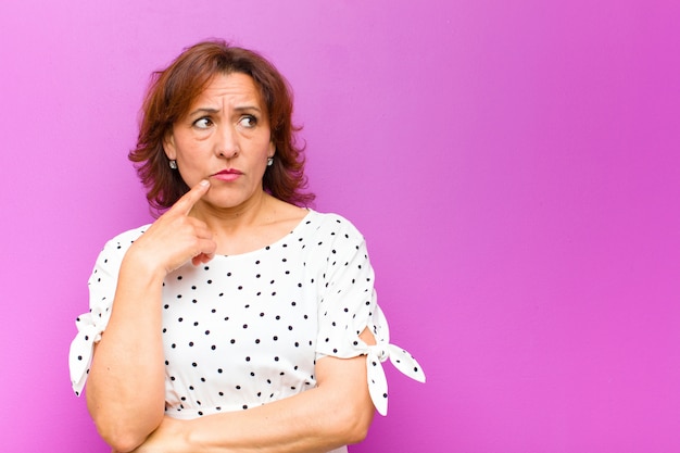 Photo middle age woman with surprised, nervous, worried or frightened look, looking to the side towards copy space against purple wall