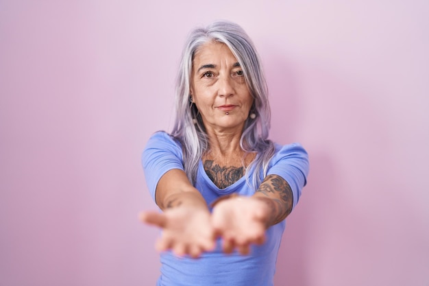 Middle age woman with tattoos standing over pink background smiling with hands palms together receiving or giving gesture hold and protection
