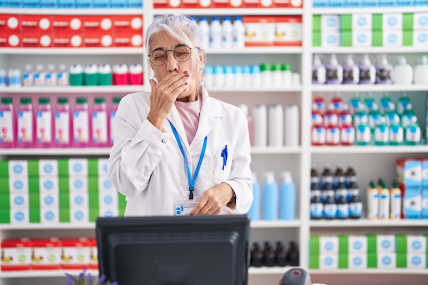 Middle age woman with tattoos working at pharmacy drugstore bored yawning tired covering mouth with hand restless and sleepiness