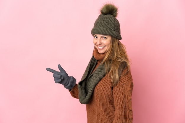 Donna di mezza età con cappello invernale isolato sul muro rosa che punta il dito a lato