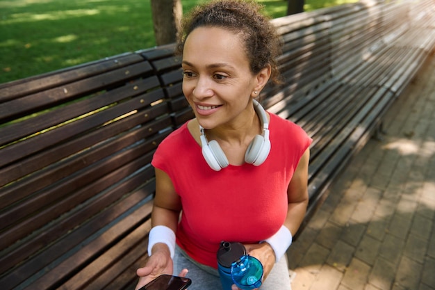 Middle aged African American fit woman athlete in bright red tshirt smiles while relaxing sitting on a wooden bench in the city park