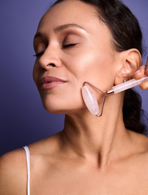 Middle aged African American pretty woman doing smoothing anti-aging face massage, massaging her cheek with pink jade stone roller massager, isolated on violet background with copy ad space. Close-up