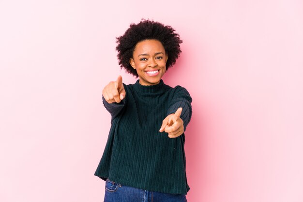 Middle aged african american woman against a pink wall cheerful smiles pointing to front.