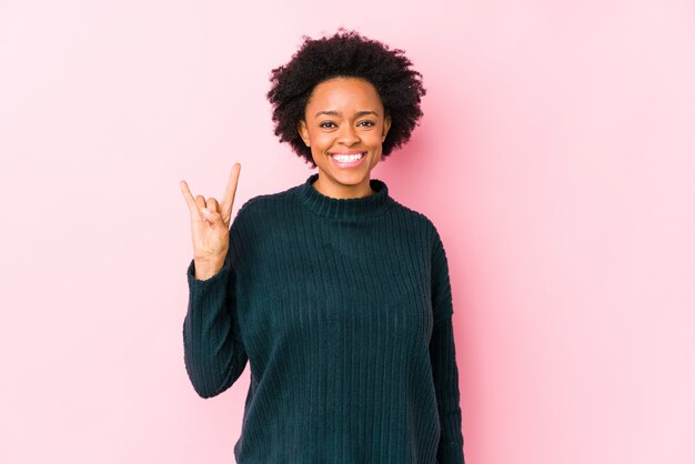 Middle aged african american woman against a pink wall isolated showing a horns gesture as a revolution concept.