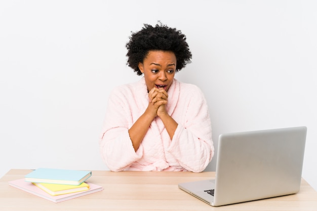 Middle aged african american woman working at home praying for luck, amazed and opening mouth looking to front.