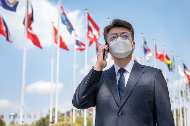 Middle-aged Asian businessman wearing a mask and using a smartphone under various national flags fluttering in the wind.