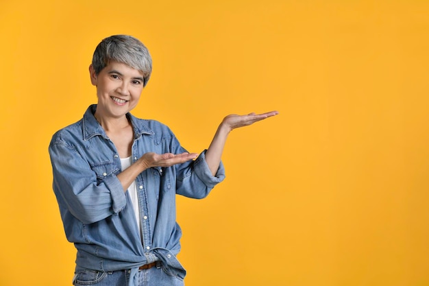 Middle aged Asian woman 50s showing empty copy space on the open hands palm for text or product isolated on colour background looking and smiling at the camerax9