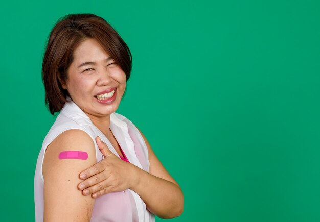 Middle aged Asian woman smiling showing her arm with bandage patch showing she got vaccinated for Covid 19 virus on green background. Concept for Covid 19 vaccination.