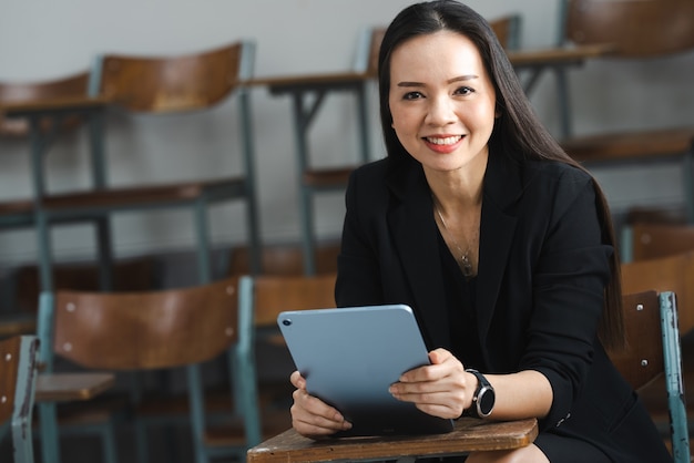 A middle-aged Asian woman teacher holding a tablet sitting in the classroom at University