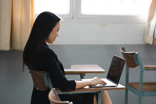 A middle-aged asian woman teacher sitting in the classroom with\
laptop on desk at university