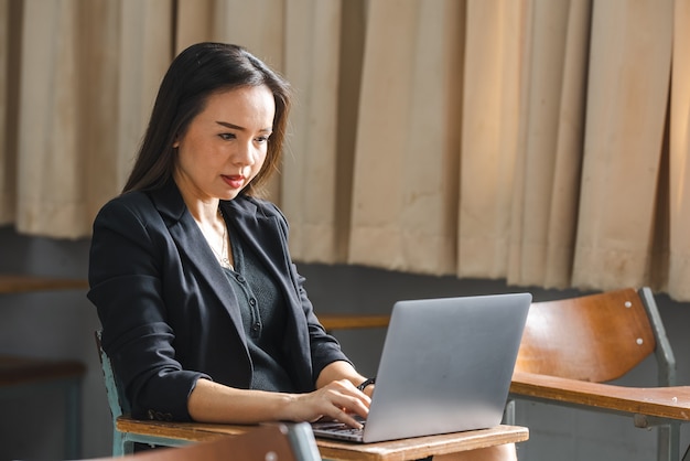 A middle-aged asian woman teacher sitting in the classroom with\
laptop on desk at university