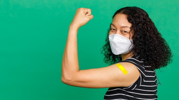 Middle aged Asian woman wearing mask showing her arm with bandage patch showing she got vaccinated for Covid 19 virus on green background. Concept for Covid 19 vaccination.