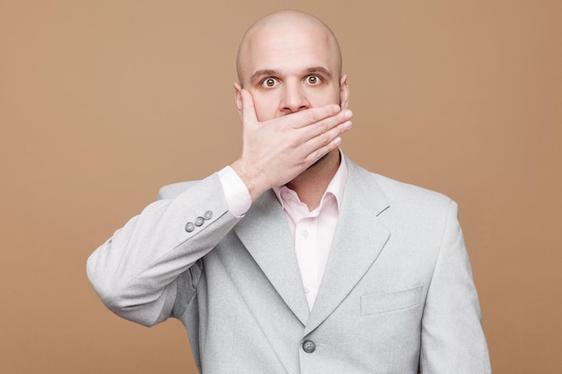 Middle aged bald bearded businessman in classic light gray suit standing and covered his mouth