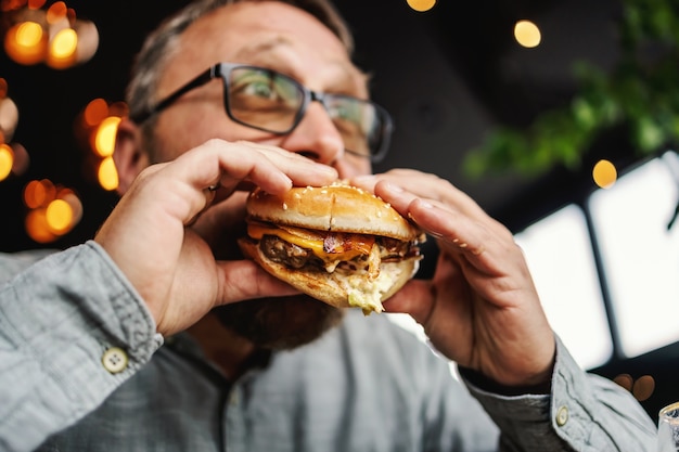 レストランに座っておいしいハンバーガーを食べている中年のひげを生やした空腹の男。