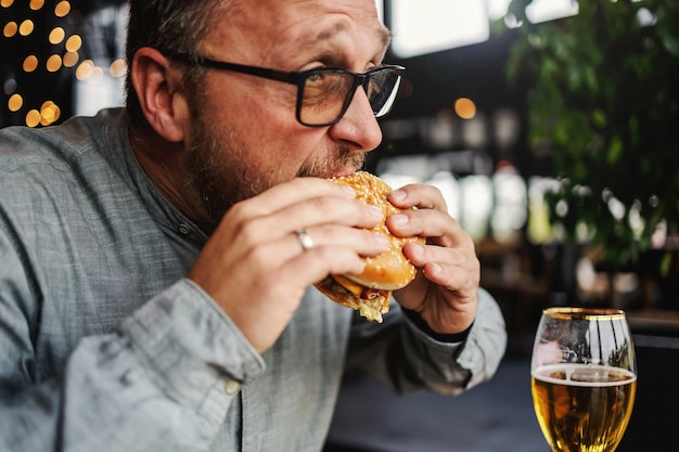 Бородатый голодный мужчина средних лет сидит в ресторане и ест вкусный гамбургер