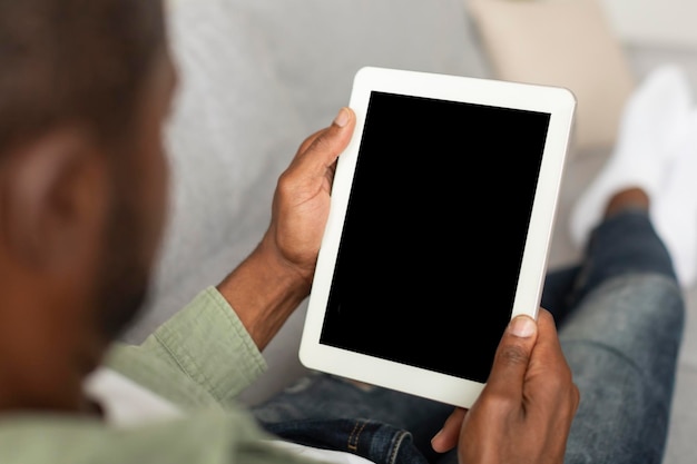 Черный мужчина средних лет смотрит на планшет с пустым экраном, используя новое приложение для работы и общения в гостиной