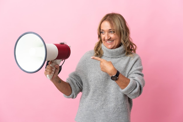 Middle aged blonde woman over isolated pink holding a megaphone and pointing side