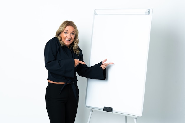 Middle aged blonde woman over isolated white background giving a presentation on white board and surprised while pointing side