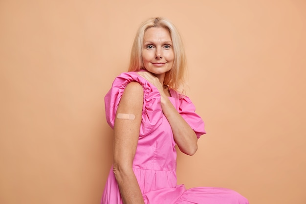 Фото Блондинка средних лет показывает руку с лейкопластырем, получает прививку от коронавируса, защищает свое здоровье во время пандемии, носит розовое платье, сидит у бежевой стены студии