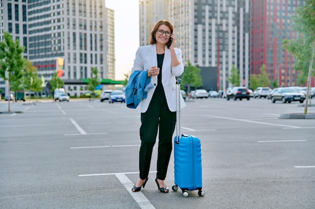 Middle aged business woman with suitcase talking on mobile\
phone in outdoor parking