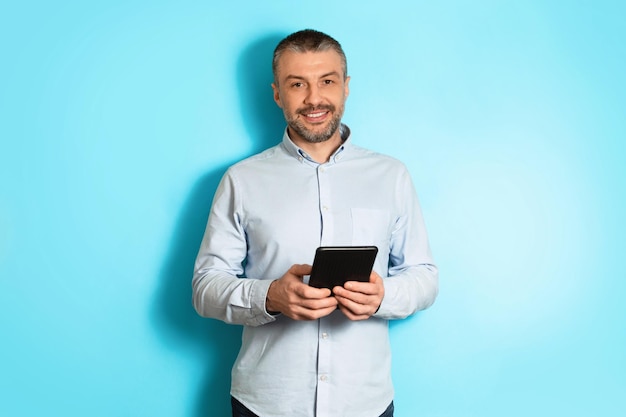 Middle aged businessman holding digital tablet standing over blue background