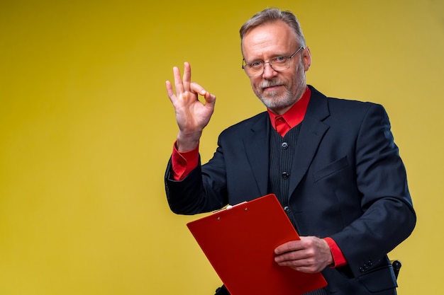 Middle aged businessman holding file folder. Showing OK sign. Horizontal format isolated on yellow background.