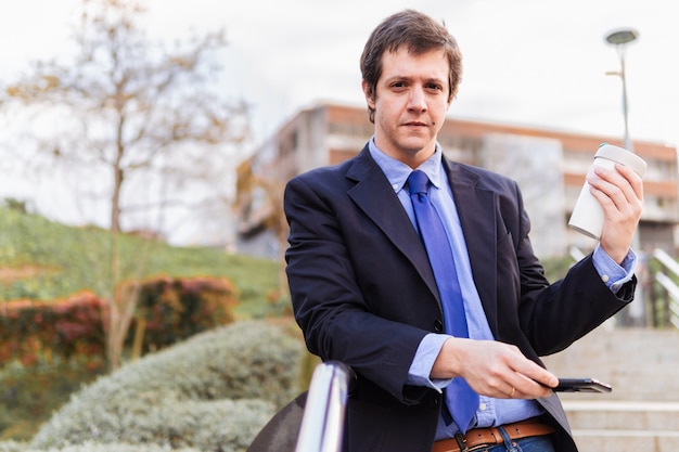 Foto uomo d'affari di mezza età in giacca e cravatta con caffè da asporto per strada in attesa di un cliente che tiene in mano uno smartphone. copia spazio