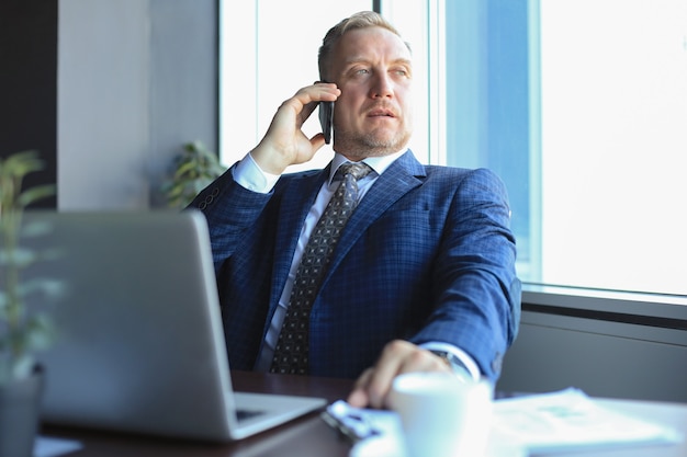 Фото Бизнесмен средних лет с ноутбуком разговаривает по телефону в современном офисе.