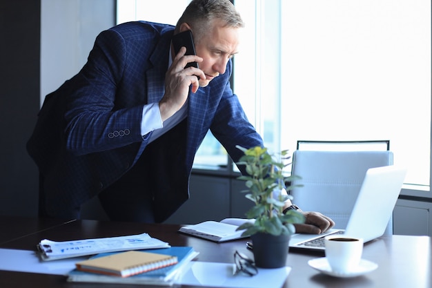 Photo middle aged businessman with laptop talking on telephone in modern office.