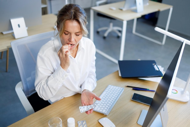 Middle aged businesswoman taking medicine pill while sitting at office workplace