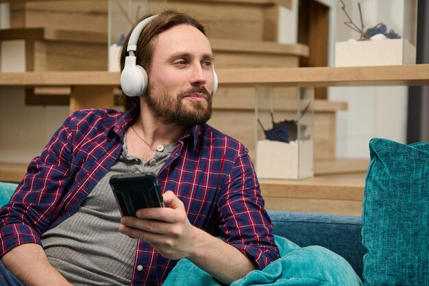 Middle aged Caucasian bearded handsome man with wireless headphones holds a smartphone in his hand and dreamily looking aside