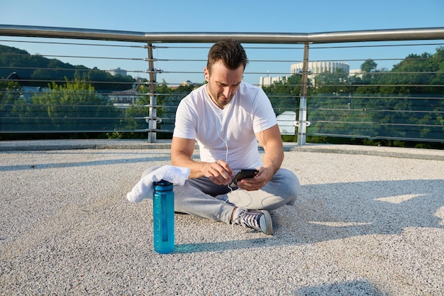 Middle aged Caucasian man athlete using fitness tracking app on smartphone Determined sportsman checking his phone sitting on the city bridge and relaxing after outdoor heavy workout