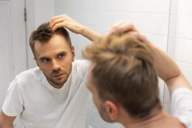 Middle aged caucasian white man with a short beard looks at his hair in the mirror in the bathroom and worried about balding The concept of the problem of male hair loss early baldness and alopecia