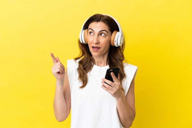 Middle aged caucasian woman isolated on yellow background listening music with a mobile and singing