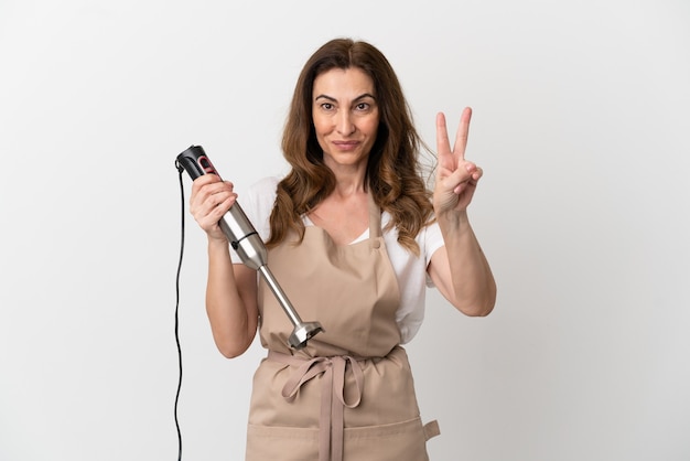 Middle aged caucasian woman using hand blender isolated on white background smiling and showing victory sign