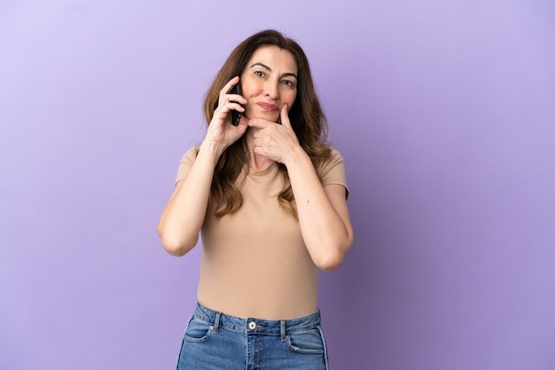 Donna caucasica di mezza età che utilizza il telefono cellulare isolato su sfondo viola felice e sorridente