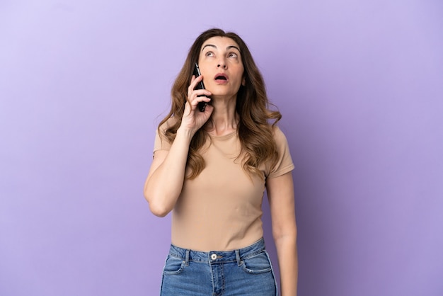 Middle aged caucasian woman using mobile phone isolated on purple background looking up and with surprised expression