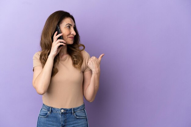 Middle aged caucasian woman using mobile phone isolated on purple background pointing to the side to present a product