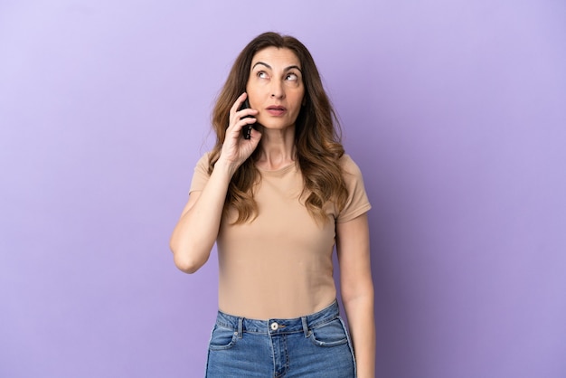 Middle aged caucasian woman using mobile phone isolated on purple background thinking an idea while looking up