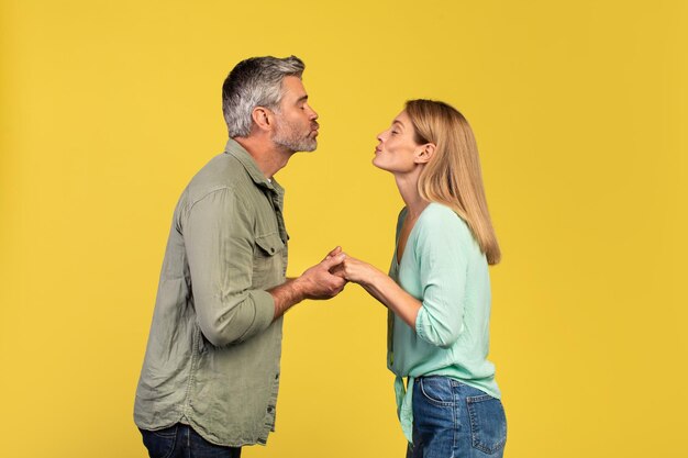 Middle aged couple in love pursing lips for kiss over yellow studio background profile side view
