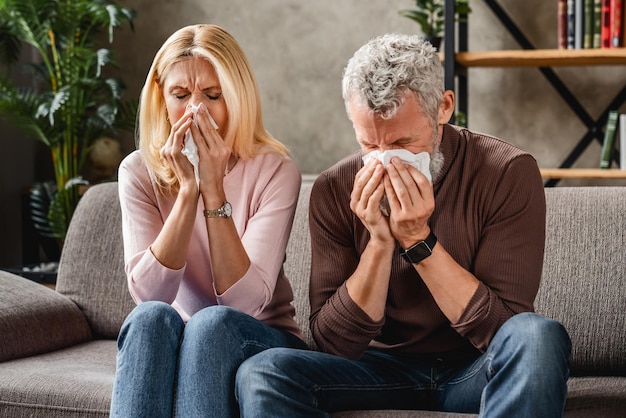 Middle aged couple sitting on sofa blowing their nose flu concept with tissues and blanket