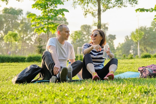 Middle-aged couple sitting on yoga mat, man and woman talking relaxing drinking water. Active healthy lifestyle, relationship, sport, fitness in mature people