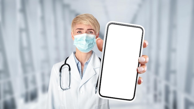 カメラで空白のスマートフォンを示す医療マスクの中年医師の女性