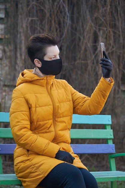 Middle aged european woman in protective black mask and gloves takes selfie on smartphone outside during coronavirus COVID-19 Epidemic